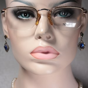 May include: A close-up of a mannequin wearing gold-rimmed glasses with clear lenses. The glasses have the word "NEW WAVE" printed on the right lens. The mannequin is also wearing blue gemstone earrings and a pearl necklace.