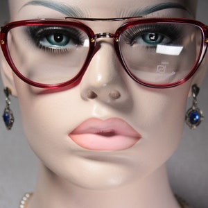 Peut inclure: Gros plan d'un mannequin portant des lunettes de soleil de forme œil de chat rouges avec des accents dorés. Les lunettes ont des verres transparents et un détail de design subtil sur la lentille droite. Le mannequin porte des boucles d'oreilles en pierres précieuses bleues.