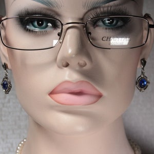 Puede incluir: Una cabeza de maniquí con gafas rectangulares con montura fina de color dorado. Las gafas tienen la marca "Chantal" impresa en la lente. El maniquí también lleva pendientes plateados con piedras preciosas azules y un collar de perlas.