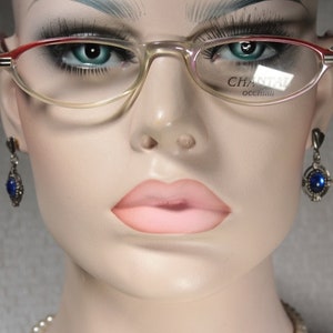 Könnte beinhalten: Eine Mannequin trägt ovale Brillen mit einem rot-weißen Rahmen. Die Gläser sind klar und auf dem rechten Glas steht der Text "CHANTAL occhiali". Die Mannequin trägt auch silberne Ohrringe mit blauen Edelsteinen.