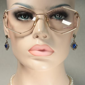 May include: Gold-rimmed eyeglasses with a geometric design. The lenses are clear, and the frame has a subtle, elegant shape. The image also shows blue gemstone earrings and a pearl necklace. The word "atrio" is printed on the lens.