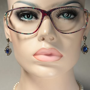 Könnte beinhalten: Nahaufnahme eines Schaufensterpuppenkopfes mit Brille und Ohrringen. Die Brille hat einen bordeaux-grünen Rahmen. Die Ohrringe sind silberfarben mit einem blauen Edelstein. Die Schaufensterpuppe hat blaue Augen und rosa Lippen.