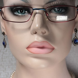 Puede incluir: Primer plano de un maniquí con gafas rojas y azules con la palabra "CHARLAL" impresa en la lente derecha. El maniquí también lleva un par de pendientes de plata con piedras preciosas azules.