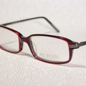 Puede incluir: Gafas rectangulares con montura burdeos y varillas negras. El nombre de la marca "PAL ZILERI" está impreso en la lente. Las gafas tienen un diseño moderno, adecuado para el uso diario.