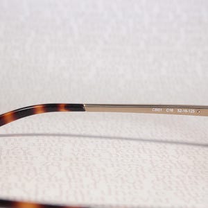 May include: Close-up of eyeglasses with a brown tortoiseshell pattern on the temples and frame. The metal arm is a light gold colour. The text "C801 C16 52-16-125" is printed on the arm.