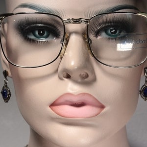 Peut inclure: Lunettes à monture dorée avec verres transparents. Les lunettes ont un design classique avec une forme rectangulaire et un pont doré. L'image montre également une paire de boucles d'oreilles en pierres bleues et un visage de mannequin.