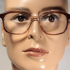 May include: A close-up of a mannequin wearing brown-framed spectacles. The spectacles have a rectangular shape and clear lenses. The mannequin's skin tone is light brown, and the background is a neutral colour.