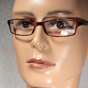 May include: Tortoiseshell-patterned, rectangular eyeglasses are shown on a mannequin. The glasses have a classic design with a price tag visible. The frame is a warm brown colour.