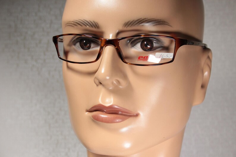 May include: Tortoiseshell-patterned, rectangular eyeglasses are shown on a mannequin. The glasses have a classic design with a price tag visible. The frame is a warm brown colour.