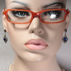 Op de afbeelding: Een mannequin draagt een oranje bril met het woord "eyewear" gedrukt op de rechterlens. De mannequin draagt ook blauwe edelsteen oorbellen.