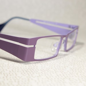 Könnte beinhalten: Eine Brille mit rechteckigem, lavendelfarbenem Gestell und modernem Design. Die Fassungen sind einfarbig lavendelfarben mit einem weißen Streifen an den Bügeln. Die Bügel sind in einem dunkleren Blaugrün gehalten.