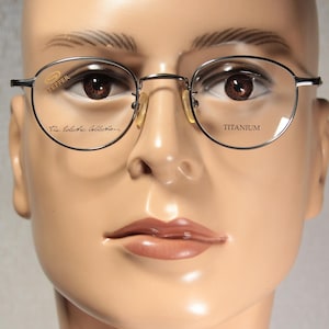 Może przedstawiać: Zbliżenie okularów ze srebrną oprawką i okrągłymi soczewkami. Okulary mają napis "PEPPER" i "The Eclectic Collection" na lewej soczewce oraz "TITANIUM" na prawej soczewce. Okulary znajdują się na manekinie.