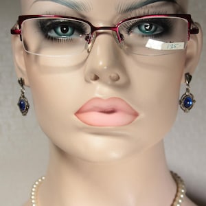 Può includere: Un manichino indossa occhiali con montatura rossa e orecchini con pietre blu. Gli occhiali hanno un design a mezza montatura. Gli orecchini sono color argento con una pietra ovale blu. Un'etichetta del prezzo è attaccata.