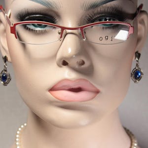 May include: A mannequin head wearing red-rimmed eyeglasses with the text "ogi" printed on the right lens. The mannequin features blue-toned eyeshadow, long eyelashes, and pink lips. It is adorned with silver-toned drop earrings featuring blue stones and a white pearl necklace.