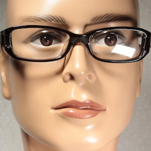 May include: Close-up of black-framed spectacles with clear lenses. The spectacles have a rectangular shape and are displayed on a mannequin head. The frame is thick and has a glossy finish.
