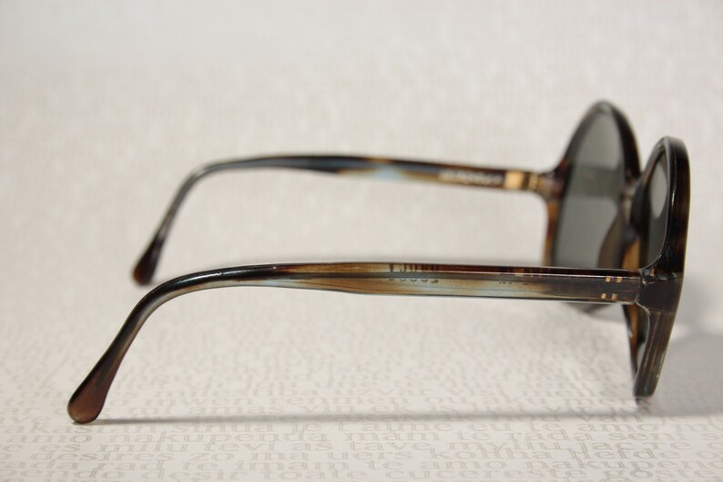May include: Brown and blue tortoiseshell framed sunglasses with round lenses. The sunglasses have a metal bar connecting the lenses and the arms. The arms are brown and blue tortoiseshell with a gold accent.