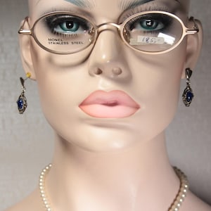 May include: A mannequin head wearing gold-rimmed oval eyeglasses with the text "MONEL STAINLESS STEEL" and a price tag. The mannequin also has blue gemstone earrings and a pearl necklace.