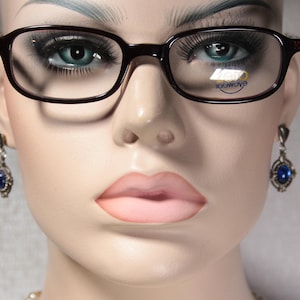 Può includere: Primo piano di un manichino che indossa occhiali marroni con lenti trasparenti e orecchini con pietre preziose blu. Gli occhiali hanno la scritta "Eyemagic" stampata sulle lenti.