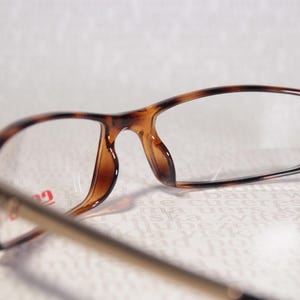 May include: Close-up of spectacles with tortoiseshell frames. The glasses have rectangular lenses and gold-coloured arms. The lenses are clear.