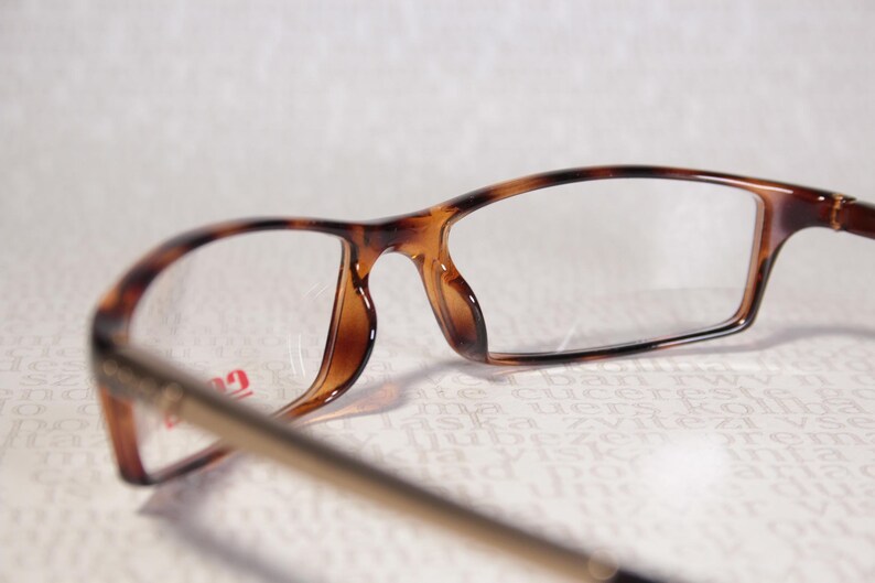 May include: Close-up of spectacles with tortoiseshell frames. The glasses have rectangular lenses and gold-coloured arms. The lenses are clear.