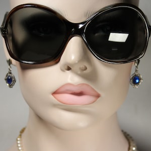 May include: A mannequin wearing large, dark brown sunglasses with a blue gemstone earring on each ear. The mannequin is wearing a pearl necklace.