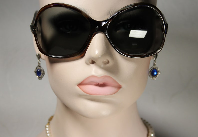 May include: A mannequin wearing large, dark brown sunglasses with a blue gemstone earring on each ear. The mannequin is wearing a pearl necklace.
