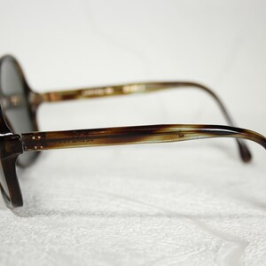 May include: Tortoise shell frame sunglasses with dark gray lenses. The sunglasses have a rounded shape and a thin metal bar connecting the lenses.