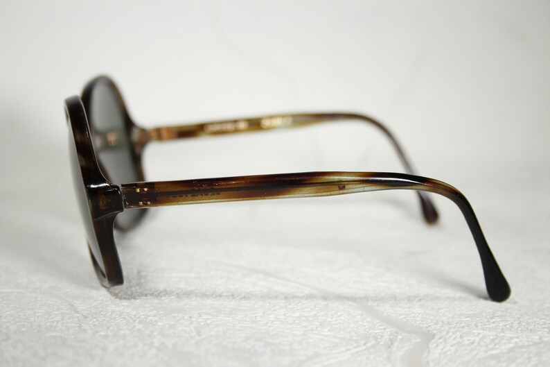 May include: Tortoise shell frame sunglasses with dark gray lenses. The sunglasses have a rounded shape and a thin metal bar connecting the lenses.