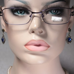 May include: A close-up of a woman's face wearing a pair of silver-rimmed glasses with purple lenses. The glasses have the word "Fontana" printed on the right lens. The woman is wearing blue gemstone earrings.