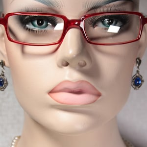 Op de afbeelding: Een close-up van een mannequin die een rode bril draagt met een blauwe edelsteen oorring. De mannequin heeft een neutrale uitdrukking en draagt een parelketting.