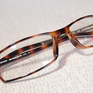 May include: Tortoiseshell-patterned spectacles with clear lenses. The rectangular frames feature the text "COM-EIGHT" on the arm. A red and white sticker is visible on the right lens.