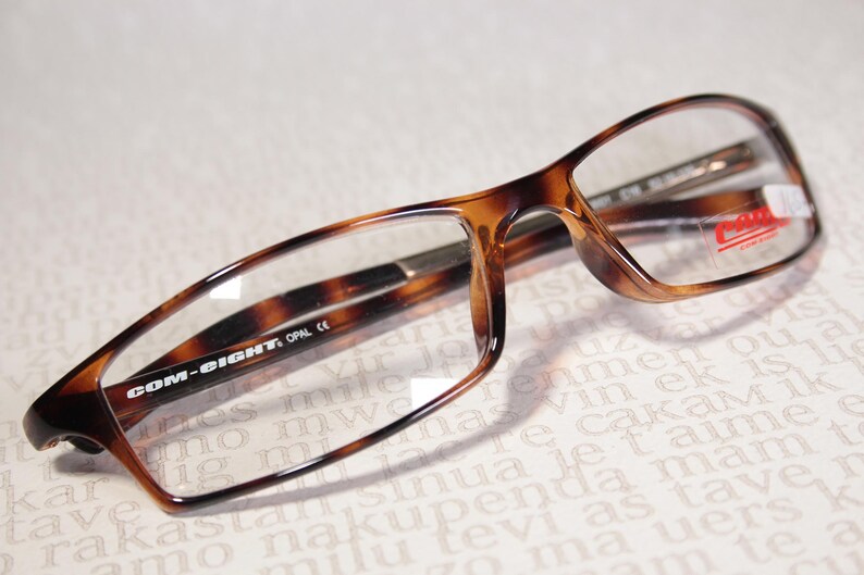 May include: Tortoiseshell-patterned spectacles with clear lenses. The rectangular frames feature the text "COM-EIGHT" on the arm. A red and white sticker is visible on the right lens.