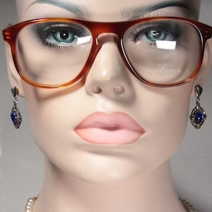 May include: A mannequin wearing brown tortoise shell eyeglasses with a blue gemstone dangle earring on each ear. The eyeglasses have a clear lens and the brand name "Eschen" is visible on the right lens.