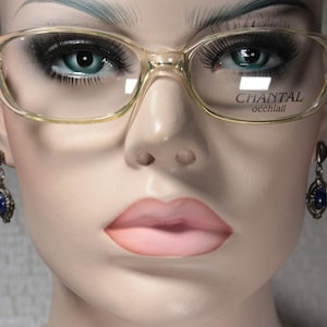 Könnte beinhalten: Eine Mannequin trägt eine klare, katzenaugenförmige Brille mit goldenen Akzenten. Die Brille hat den Text "CHANTAL occhiali" auf der rechten Linse.