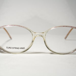 Pode incluir: Óculos ovais de plástico transparente com aro rosa e o texto "EURO SPRING HINGE" impresso na armação.