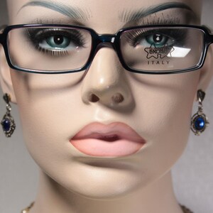 Op de afbeelding: Een close-up van een vrouwengezicht met een zwarte bril met de tekst "Luxottica Italy" op de rechterlens. Ze draagt blauwe edelsteen oorbellen.