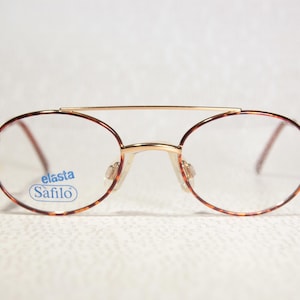 May include: Vintage spectacles with a gold-tone metal frame and oval lenses. The rims feature a tortoiseshell pattern. The brand name "elasta Safilo" is visible on one lens. The glasses have a double bridge design.