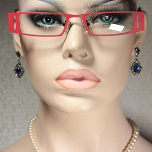 Pode incluir: Uma mulher usando óculos de armação vermelha com brincos de pedra preciosa azul. Os óculos têm uma armação vermelha fina e os brincos são de prata com uma pedra preciosa azul.