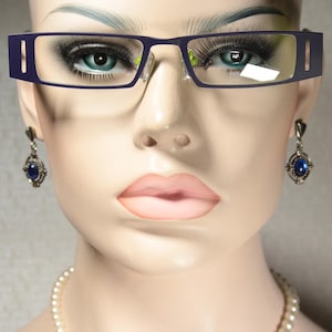 Könnte beinhalten: Eine Mannequin trägt eine dunkelblaue Brille mit einem dünnen gelben Streifen auf der Nasenbrücke. Die Mannequin trägt auch silberne Ohrringe mit blauen Edelsteinen.