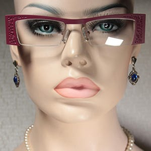 Puede incluir: Primer plano de la cara de una mujer que lleva unas gafas de color burdeos con un diseño decorativo. Las gafas tienen lentes transparentes. La mujer también lleva un par de pendientes de plata con piedras preciosas azules.