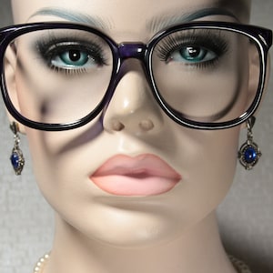 May include: A mannequin head wearing large, square-framed spectacles with a dark purple frame. The mannequin has blue eyes, long eyelashes, and pink lips. Dangling earrings with blue gemstones are visible.