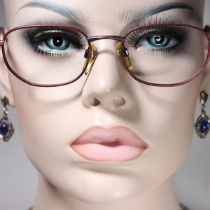 Könnte beinhalten: Nahaufnahme eines Mannequins, das eine rot umrandete Brille mit goldenen Akzenten trägt. Das Mannequin trägt blaue Edelsteinohrringe.