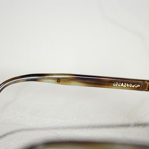 May include: Close-up of the brown tortoise shell frame of a pair of sunglasses. The frame has a gold metal hinge and the text "02CA240" is printed on the frame.