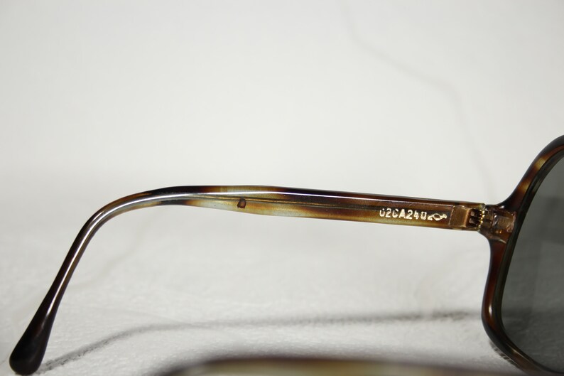 May include: Close-up of the brown tortoise shell frame of a pair of sunglasses. The frame has a gold metal hinge and the text "02CA240" is printed on the frame.