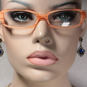 Op de afbeelding: Een mannequin draagt een paar oranje gestreepte brillen met heldere glazen. De bril heeft de tekst "HILLSIDE OPTIC" op het rechter glas.