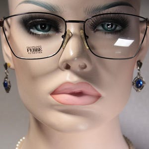 Könnte beinhalten: Schwarz gerahmte Brille mit quadratischer Form und goldenen Akzenten. Die Brille trägt den Text "GIANFRANCO FERRE LUNETTES" auf dem Glas. Die Schaufensterpuppe trägt auch blaue Edelsteinohrringe.