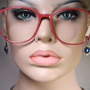 May include: A mannequin head wearing large, red-framed spectacles. The spectacles have a square shape. The mannequin also has blue-gemstone earrings and full, pink lips. The background is a gradient of light and dark grey.