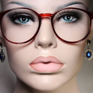 May include: A close-up of a mannequin wearing red round glasses with clear lenses. The mannequin is wearing blue earrings with a dark blue stone.