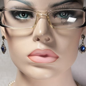 Può includere: Primo piano di un manichino che indossa occhiali con montatura trasparente e aste nere. Gli occhiali hanno la scritta "Studio One Eyewear" stampata sulla lente destra. Il manichino indossa orecchini con pietre preziose blu.