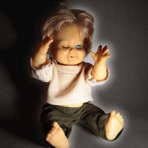 Peut inclure: Une poupée vintage aux cheveux blonds, un œil bleu et un œil fermé. La poupée porte une chemise blanche avec une bordure bleue et un pantalon vert olive. Elle est posée avec les bras levés et les pieds tendus.